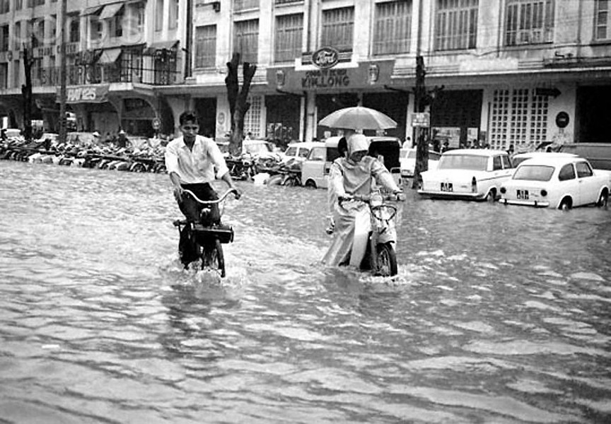 Một người đàn ông đạp xe và hai người phụ nữ lái xe máy đi trên đại lộ Lê Lợi, lúc này đang ngập chìm trong nước do mưa lớn,  Sài Gòn cuối tháng 5 năm 1968. Ảnh: Eddie Adams.