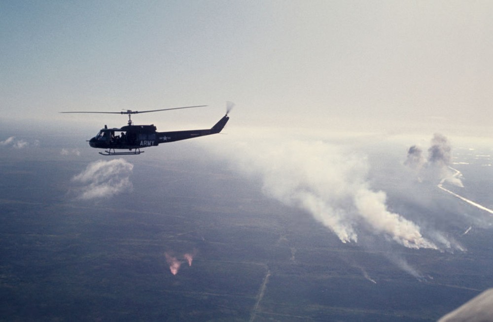 Một chiếc trực thăng UH-1 của Mỹ bay qua vùng nông thôn miền Nam thời  chiến tranh Việt Nam, năm 1965. Dưới mặt đất, khói bốc lên từ những khu vực bị quân Mỹ oanh tạc. Ảnh: Jean-Claude Sauer.