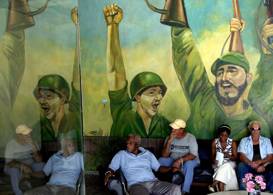 Người dân Cuba ngồi phía trước bức vẽ graffiti khắc họa chân dung lãnh tụ Cuba Fidel Castro và em trai Raul trên bức tường ở phòng chờ của văn phòng Đảng Cộng Sản Cuba.