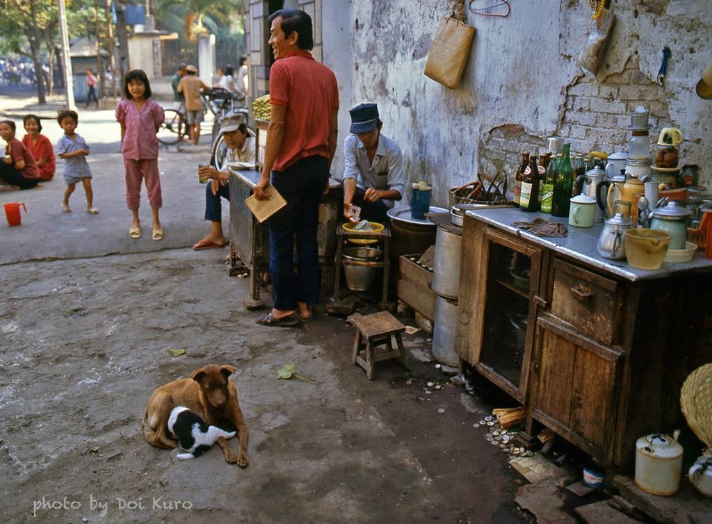 Cảnh đời thường lý thú tại một quán  cà phê vỉa hè Sài Gòn năm 1989. Chiếc tủ cũ kỹ chất đủ thứ chai lọ, ở vị trí cao nhất là một phin cà phê. Cạnh đó, chó con rúc vào bụng chó mẹ để bú sữa. Ảnh: Doi kuro/ Facebook.