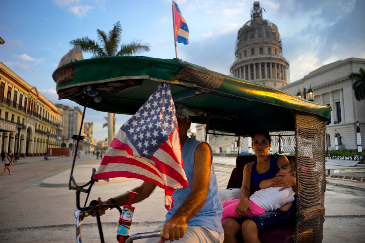 Một anh tài xế xe chở khách thông dụng ở thủ đô La Habana treo cờ Mỹ đang đi ngang qua tòa nhà El Capitolio được làm giống với tòa Capitol ở Washington. Trong hơn một năm qua,  quan hệ Mỹ-Cuba đã được cải thiện đáng kể sau những động thái mang tính xây dựng. Ảnh USN